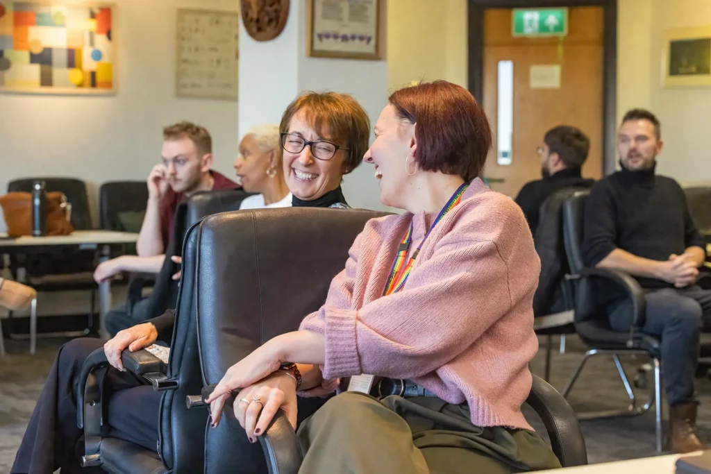 Two women laughing together while seated at an indoor gathering, with other attendees visible in the background