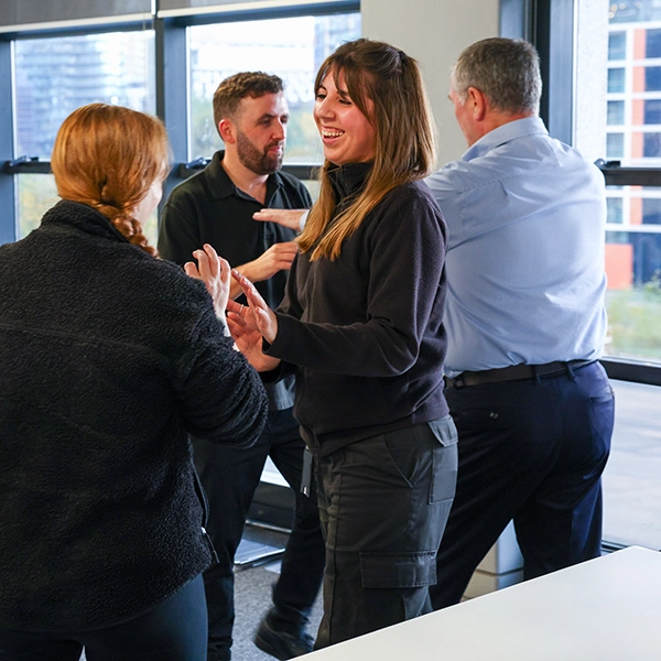 Four colleagues engaged in an animated conversation in a modern office, with one woman smiling and gesturing expressively