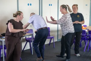 Three adults practicing a physical technique in a classroom, with a fourth person observing and smiling nearby