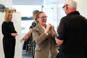 A woman in a blazer laughs enthusiastically while interacting with colleagues during an office workshop or team activity