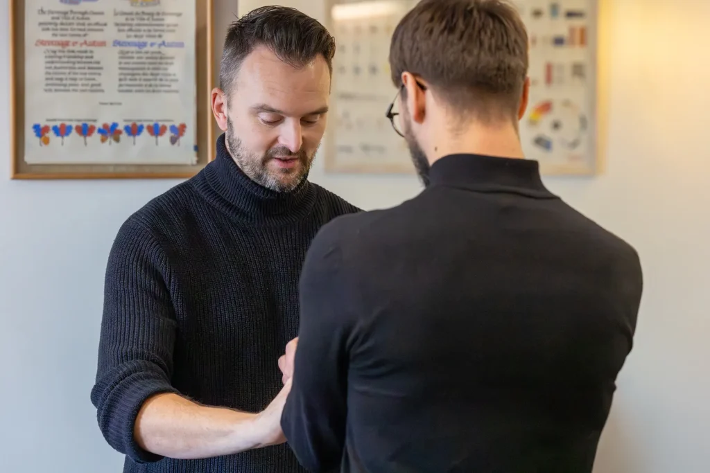 A practitioner examines a patient's arm in a clinical setting with framed certificates on the wall behind them
