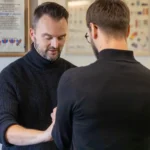 A practitioner examines a patient's arm in a clinical setting with framed certificates on the wall behind them
