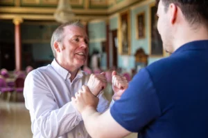 Two men holding hands and dancing together in an ornate ballroom with gilded paintings and pink chairs