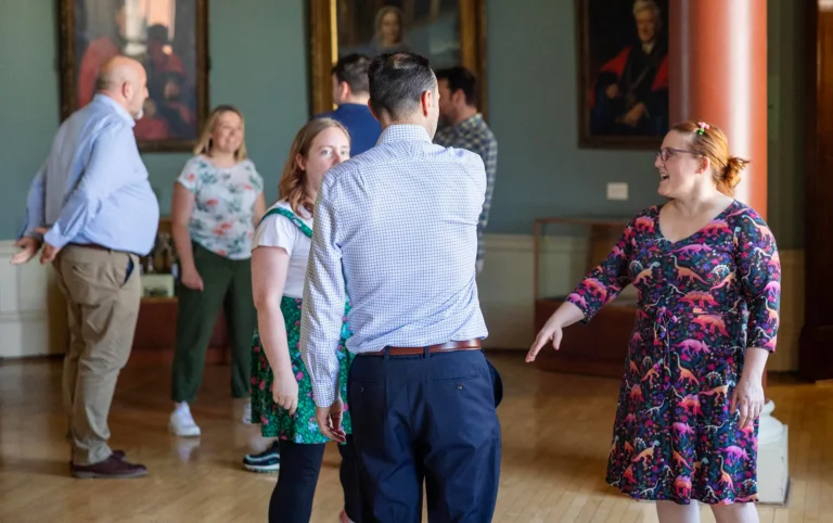 A group of people dancing in a grand hall with portrait paintings on the walls and hardwood floors