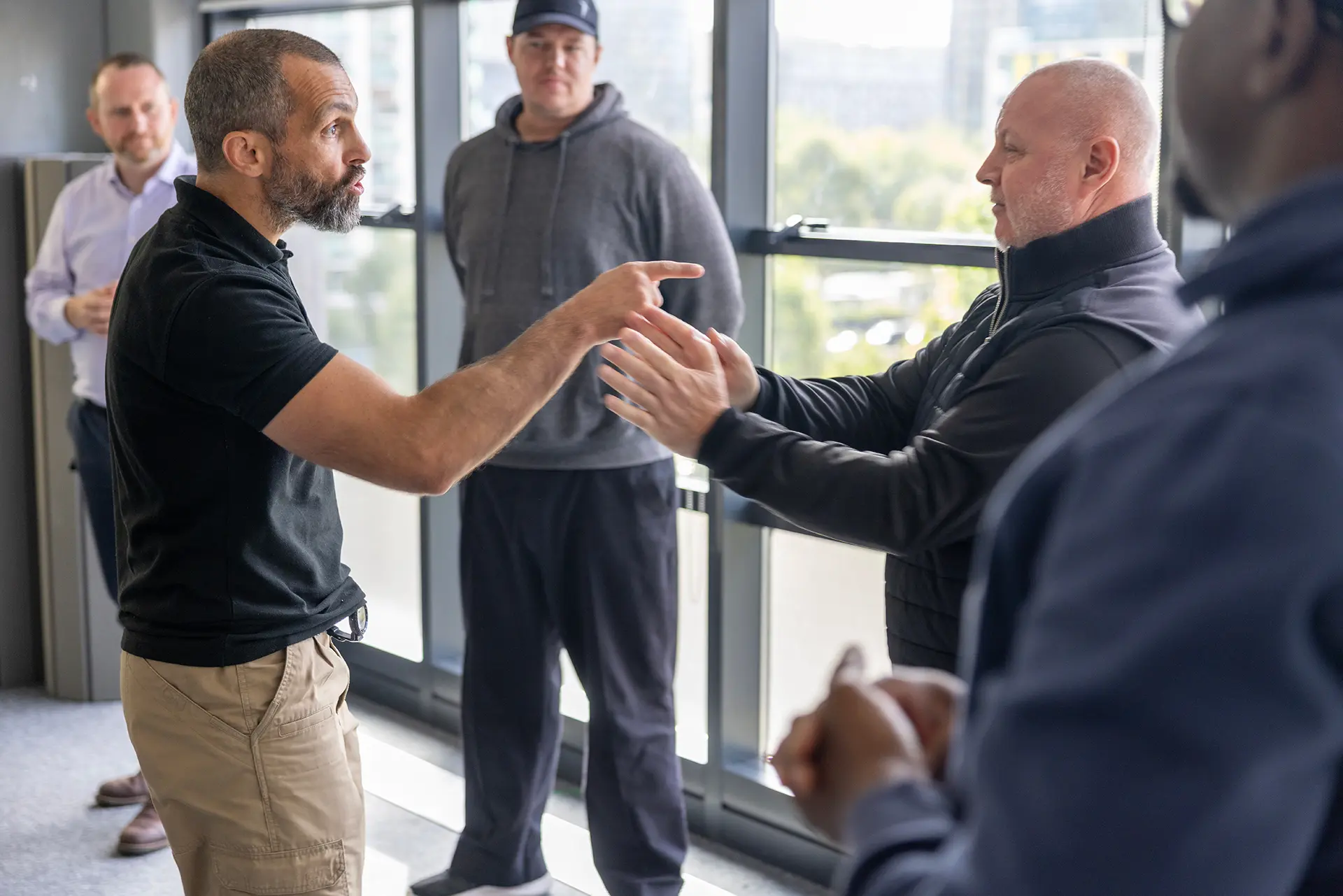 Two men face each other in an animated discussion with gesturing hands while others look on in an indoor setting