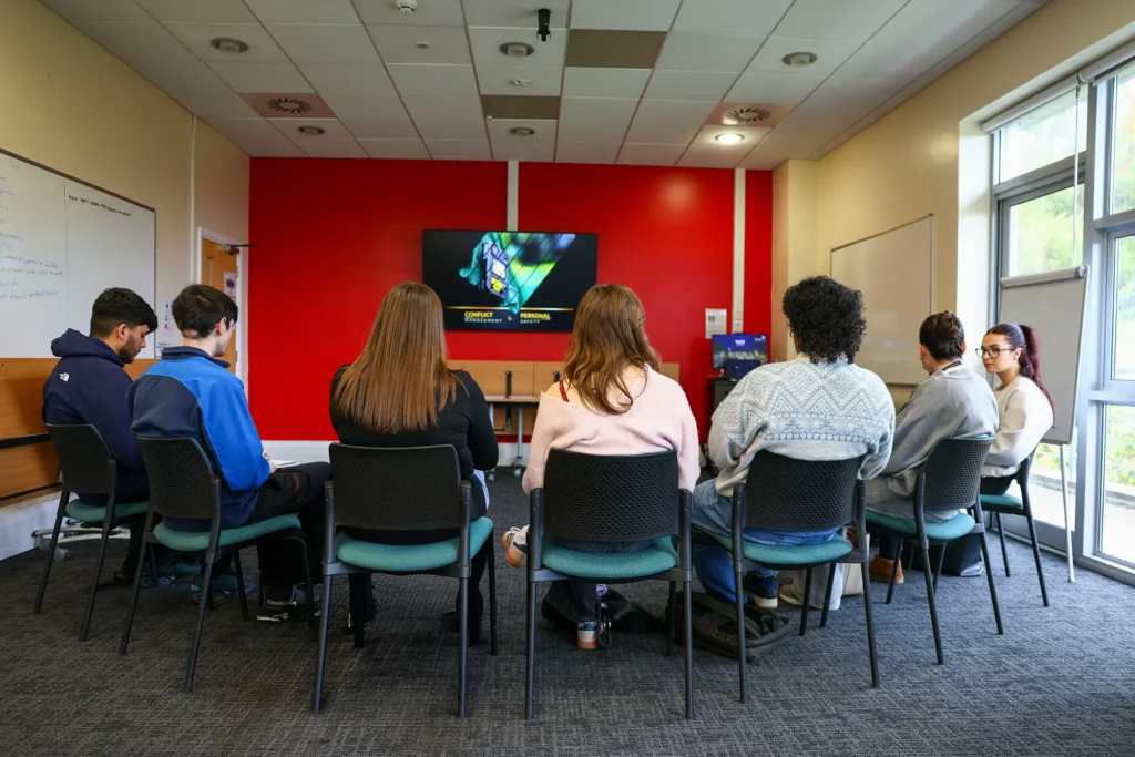 Students seated in a semicircle watching a Conflict Management and Personal Safety presentation on a wall-mounted screen