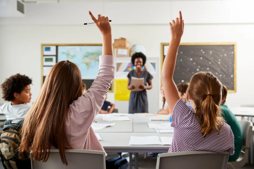 Students raise their hands to participate in a classroom lesson led by their teacher