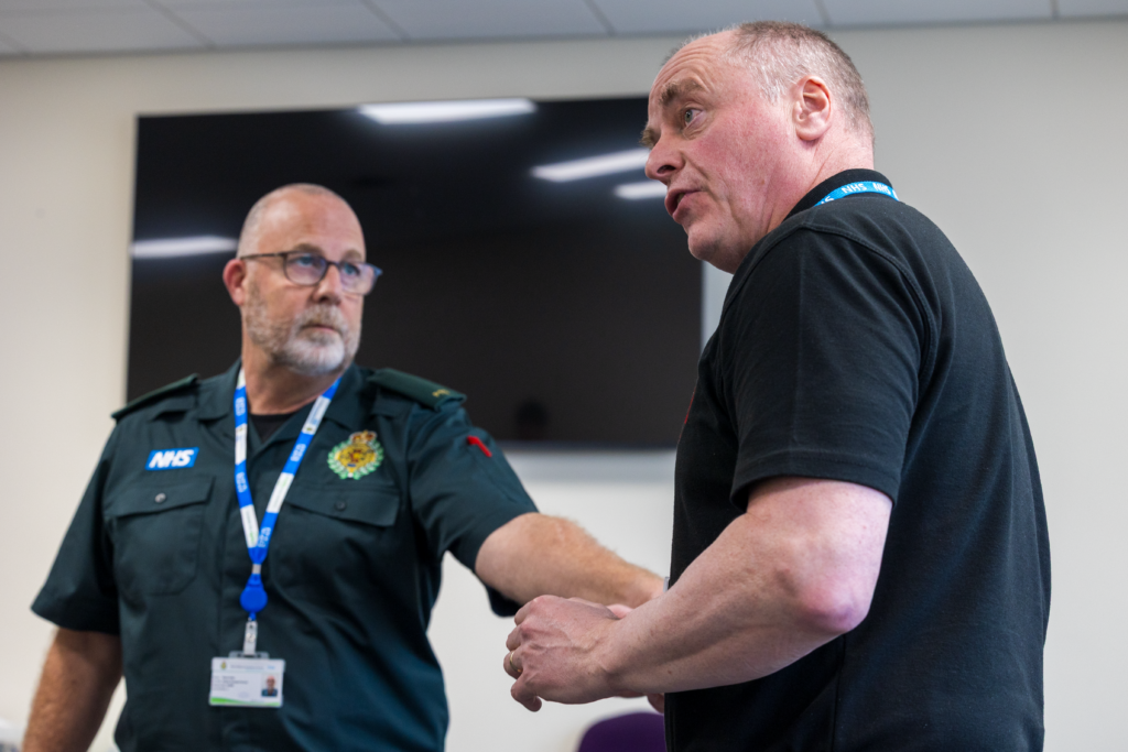 Two men in a meeting room, one wearing an NHS ambulance service uniform with a lanyard, the other speaking and gesturing
