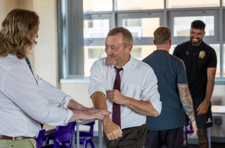 A man in a white shirt and tie practices a martial arts or self-defense move with another person indoors, as two others watch and smile in the background