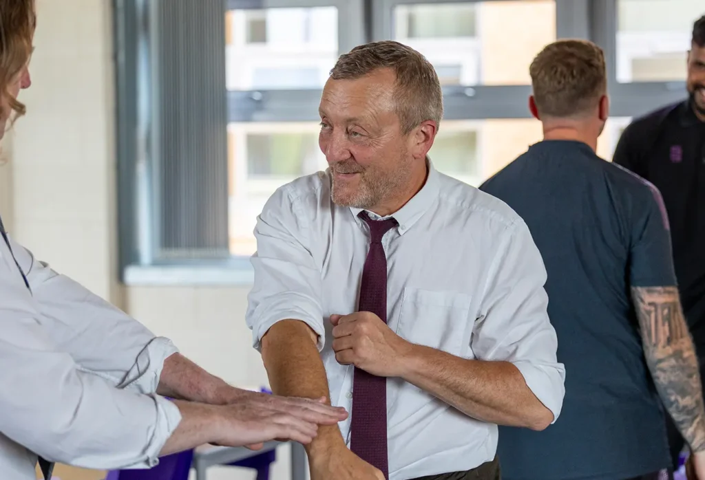 Man in a white shirt and burgundy tie smiling while greeting someone at an indoor event