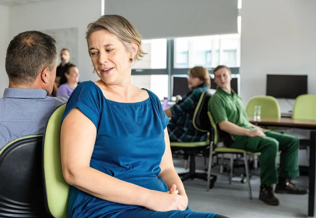 A woman in a blue top speaks with a colleague in a modern office, with other coworkers conversing in the background