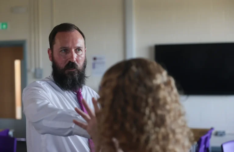A bearded man in a white lab coat and pink tie speaks and gestures to a person with curly hair in a clinical setting