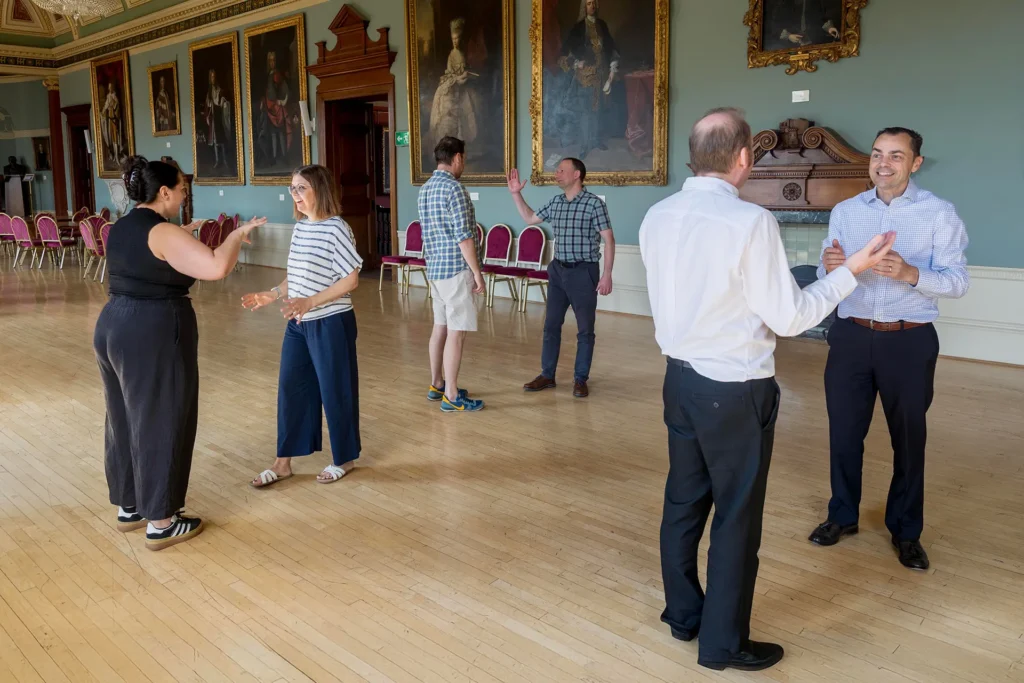 Six people engaged in a group activity or discussion in an ornate hall with large portrait paintings and a wooden floor