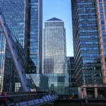 Canary Wharf financial district skyline in London, with One Canada Square tower centered among glass skyscrapers and a pedestrian bridge in the foreground