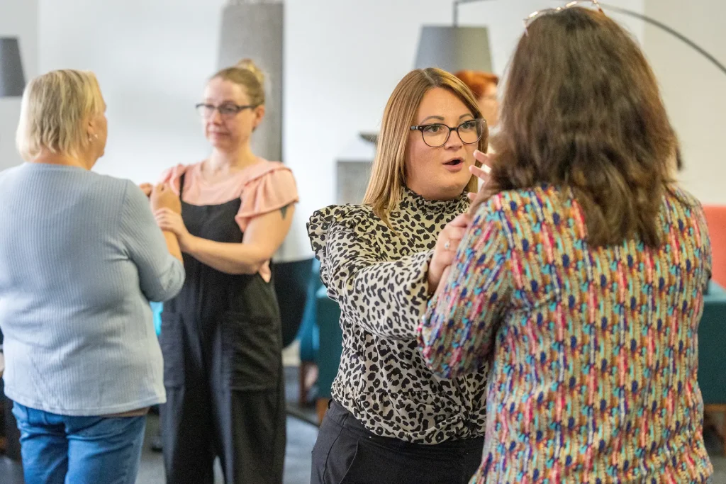 A woman in a leopard-print top gestures while speaking to another person, as two other women interact in the background indoors