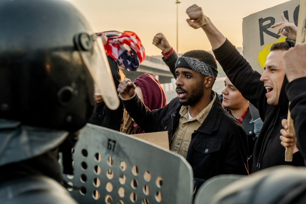 Group of disgruntled young multi-ethnic people raising arms and screaming protecting speech at riot while standing against police with shields