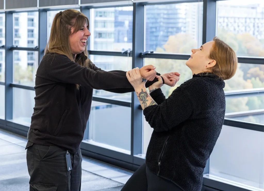 Two women practice self-defense moves together, laughing, in a bright indoor space with large windows overlooking a city