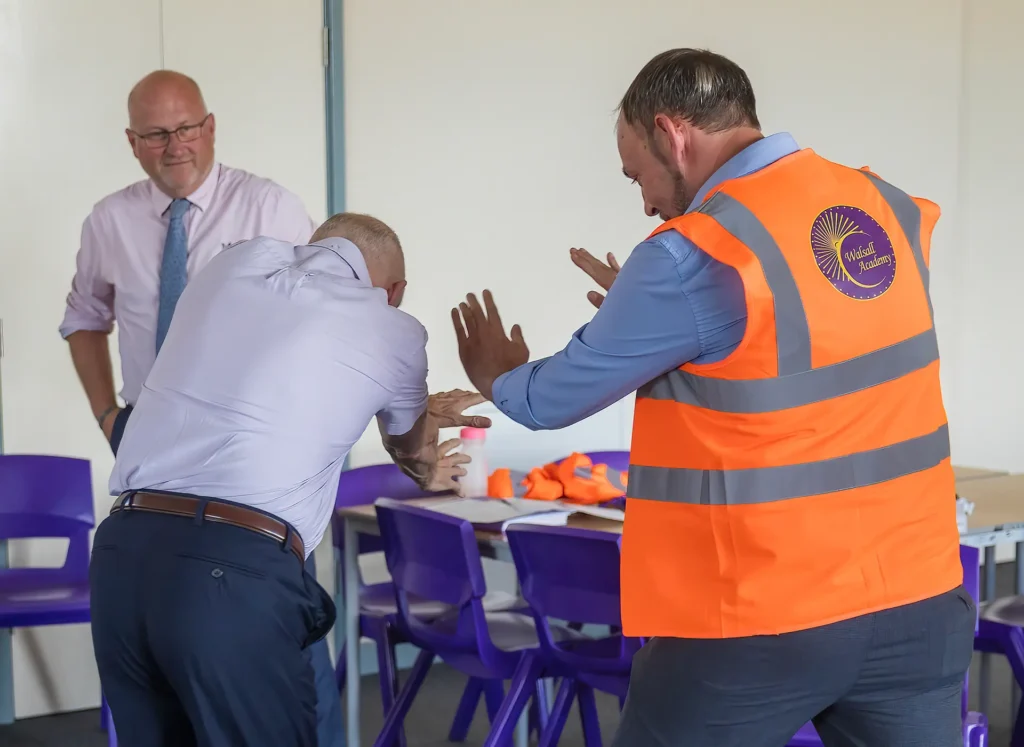 A Walsall Academy staff member in an orange hi-vis vest demonstrates a first aid technique on a man while another man looks on in a classroom