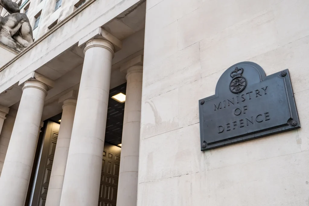 Exterior of the UK Ministry of Defence building in London, with a metal plaque reading "Ministry of Defence" mounted on a stone wall beside large columns