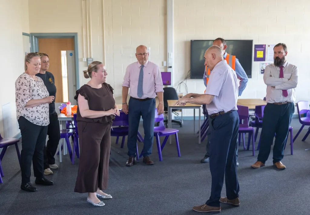 A man addresses a small group of adults standing in a school classroom with purple chairs and tables