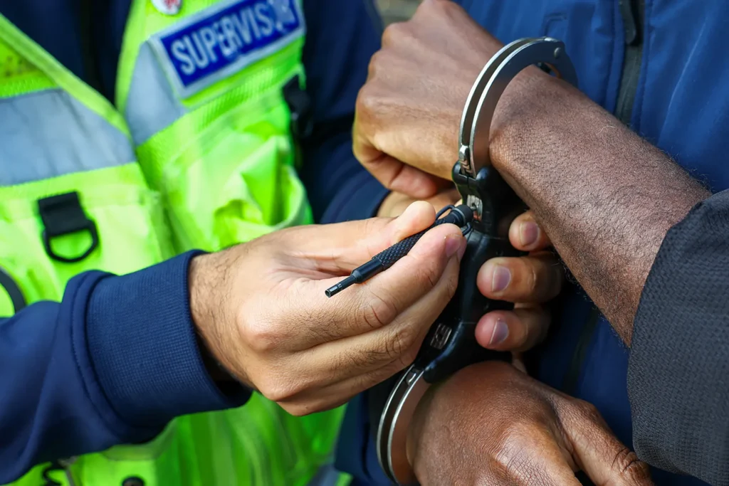 Officer using a key to unlock handcuffs on a person's wrists, with a supervisor in a high-visibility vest nearby