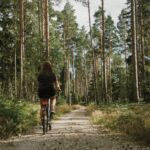 A woman riding a bicycle away from the camera along a gravel path through a tall pine forest