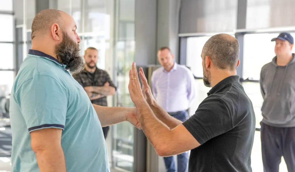 Two men face each other during a hands-on training exercise while three others observe in the background indoors